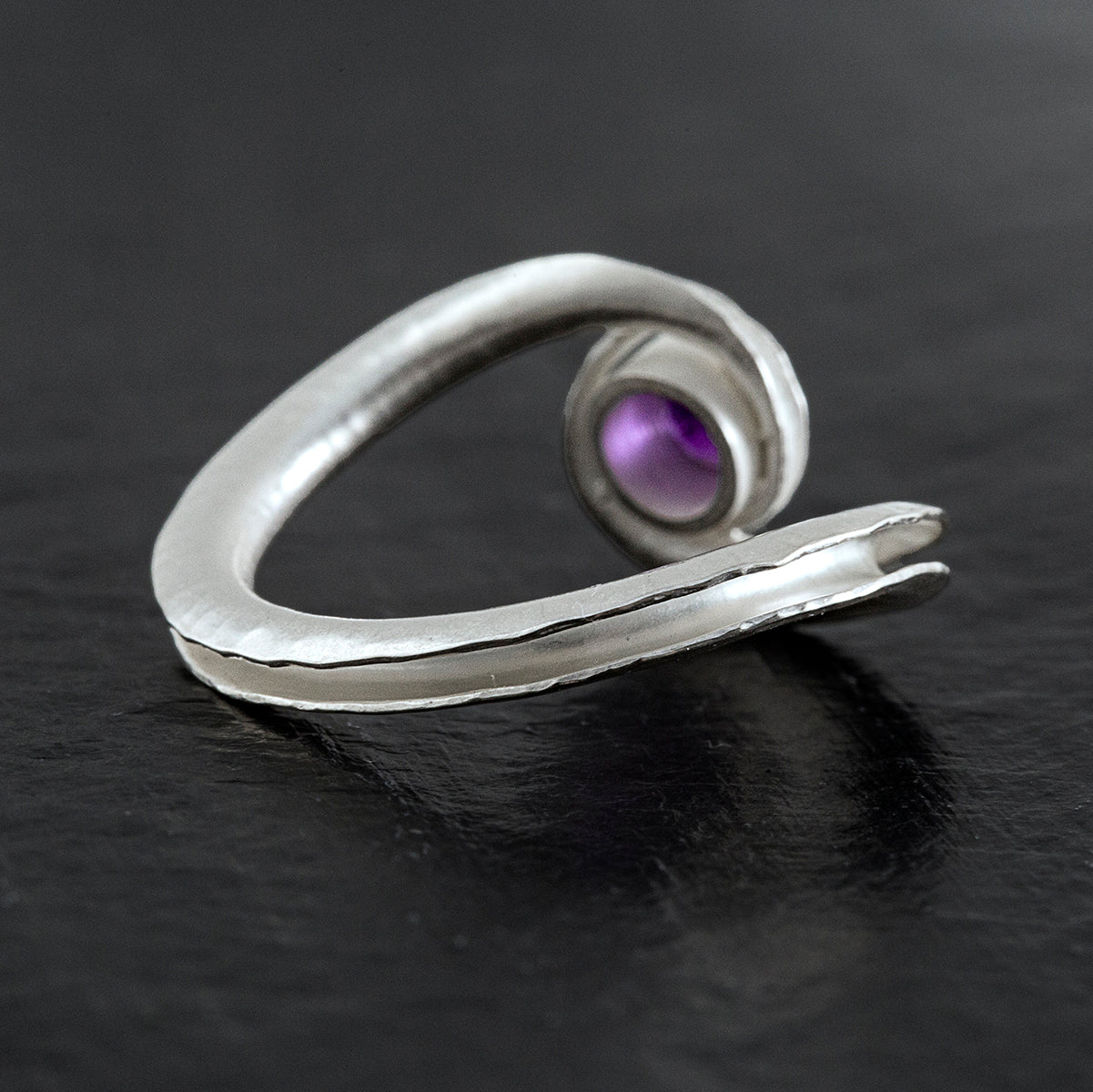 A silver birthstone ring handmade from recycled sterling silver, with a gemstone cradled between two curling ends. Shown here is the amethyst variant for February, from the back and side.