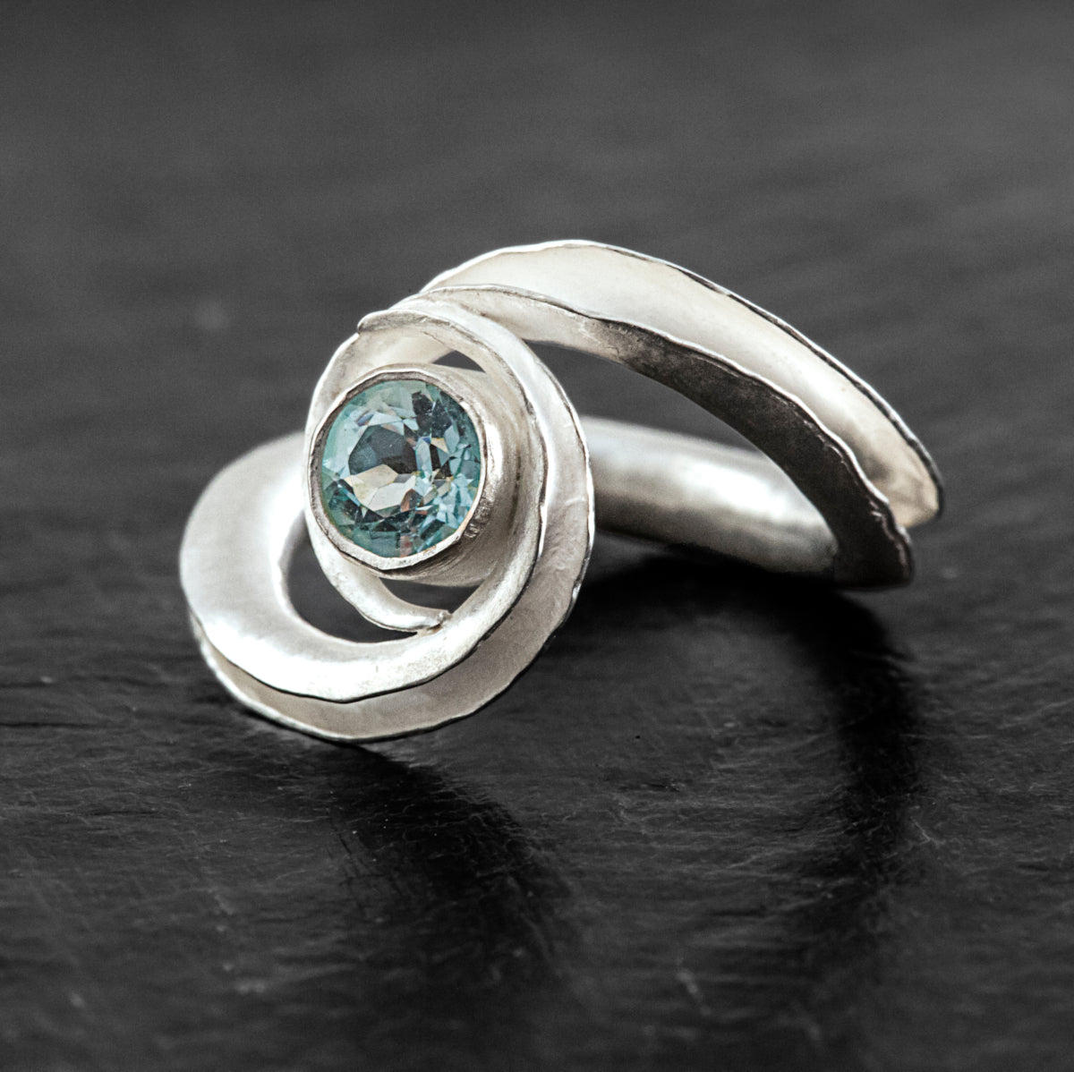 A silver birthstone ring handmade from recycled sterling silver, with a gemstone cradled between two curling ends. Shown here is the pale blue topaz variant for December or November, from the front and side.