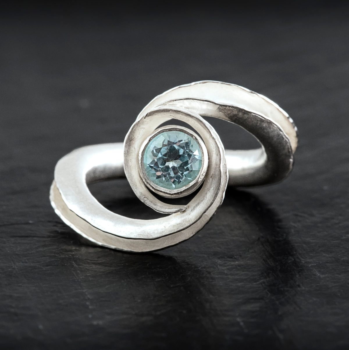 A silver birthstone ring handmade from recycled sterling silver, with a gemstone cradled between two curling ends. Shown here is the pale blue topaz variant for December or November, from the front.