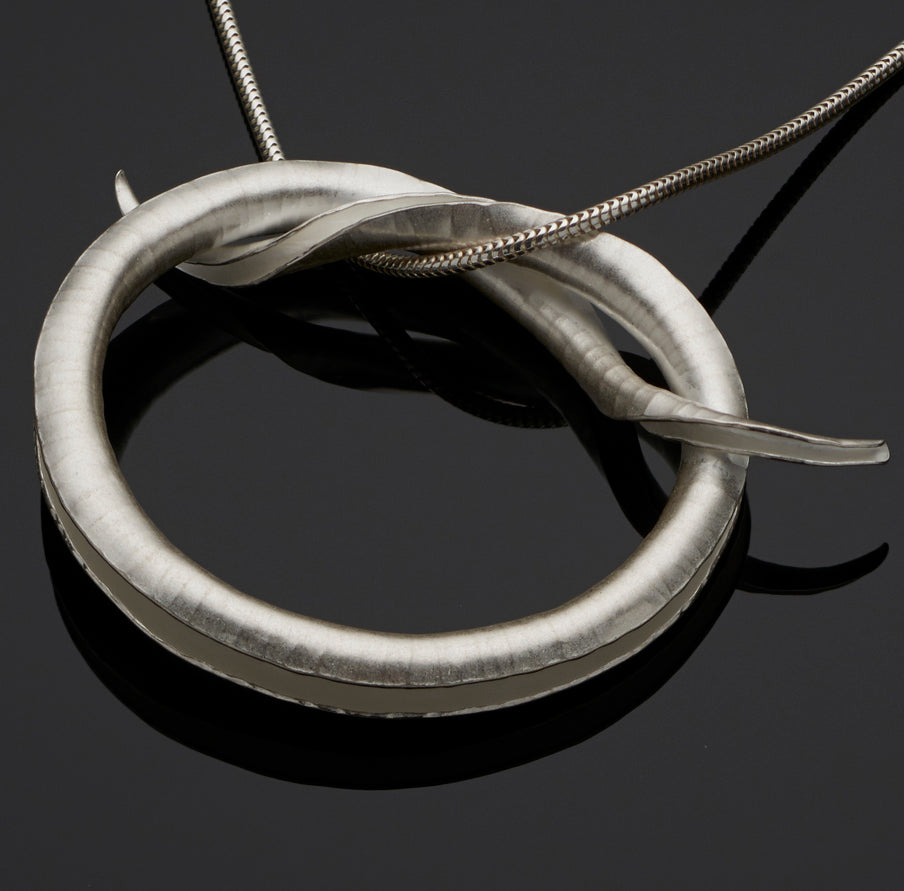 A silver pendant in the shape of a knot, in the form of a tube open down one side, hanging from a silver snake chain.