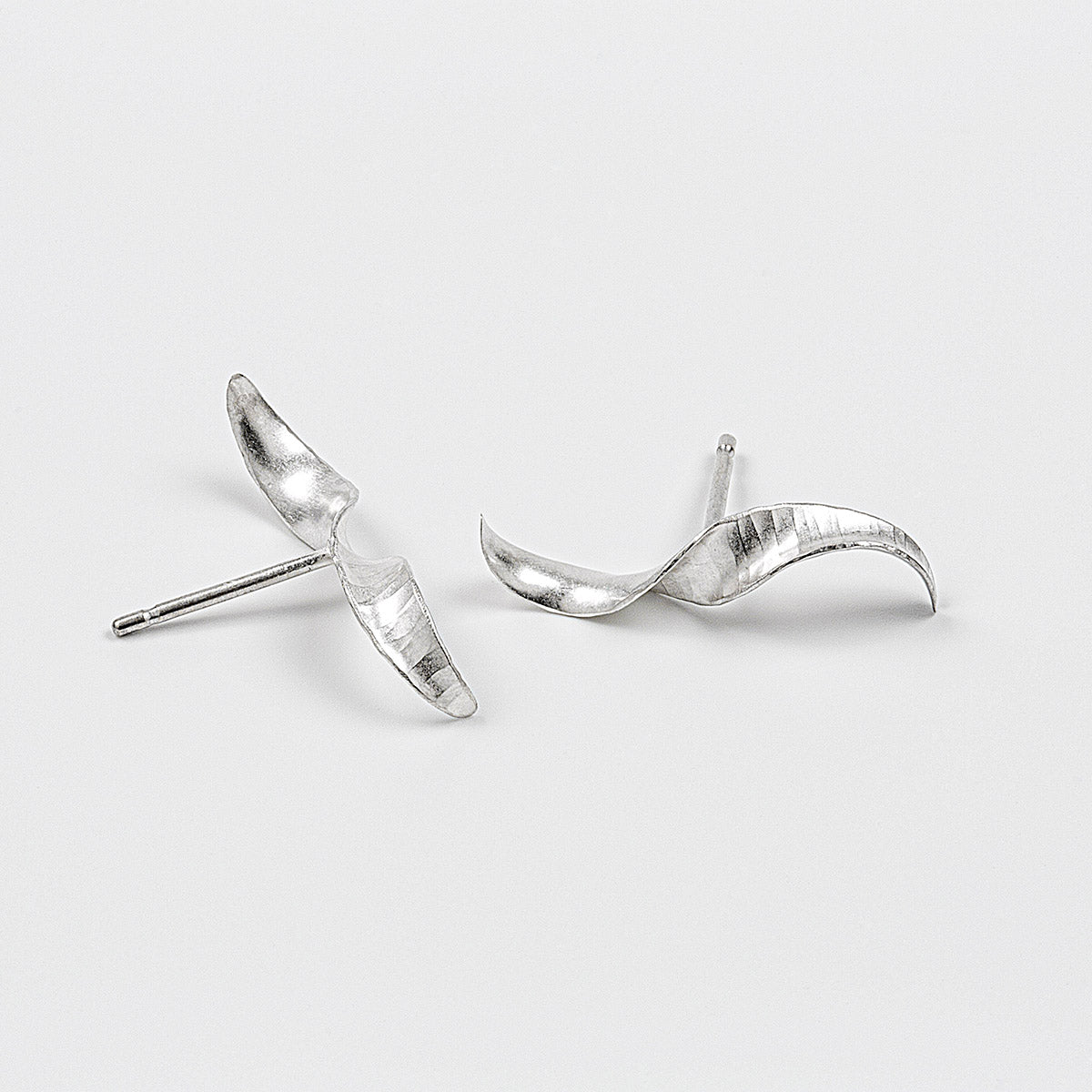 A pair of simple mirror-image silver twist earrings. They each have a stretched-S shape, a hammered texture, non-shiny finish and glittering edges. Shown on a white background, with the two at different angles.