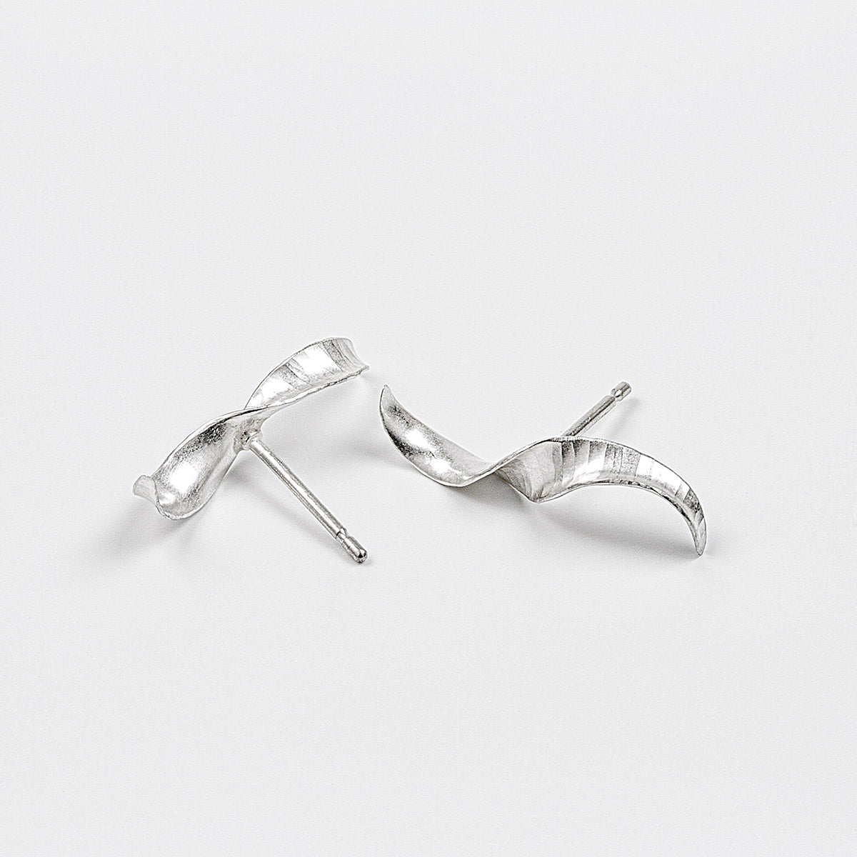 A pair of simple mirror-image silver twist earrings. They each have a stretched-S shape, a hammered texture, non-shiny finish and glittering edges. Shown on a white background, with the two at different angles.